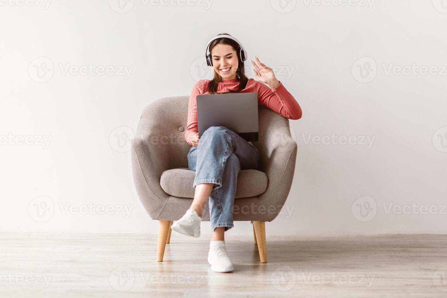 A young brunette woman sits comfortably in a light gray armchair, wearing headphones and smiling while using her laptop. She engages in an enjoyable online activity in a bright, minimalistic space. photo