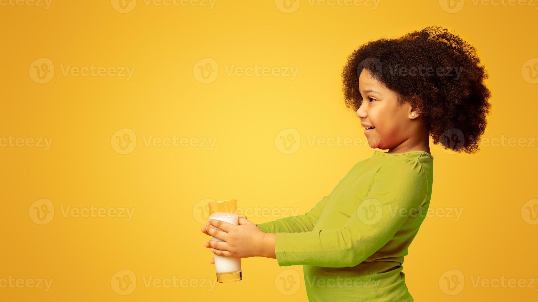 Side View Portrait Of Adorable Little Black Girl Holding Glass With Milk And Offering It, Standing On Yellow Background In Studio, Free Space photo