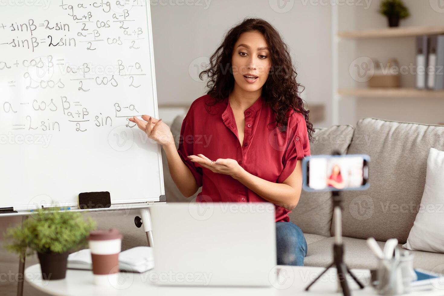 A young Middle Eastern woman teaches a math lesson remotely from her cozy living room. She engages with students through a laptop while referencing notes on a whiteboard. photo