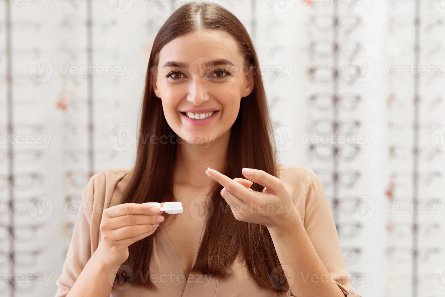 A smiling woman holds a contact lens case and applies solution with care. The setting is bright and the background features various eyeglasses. photo
