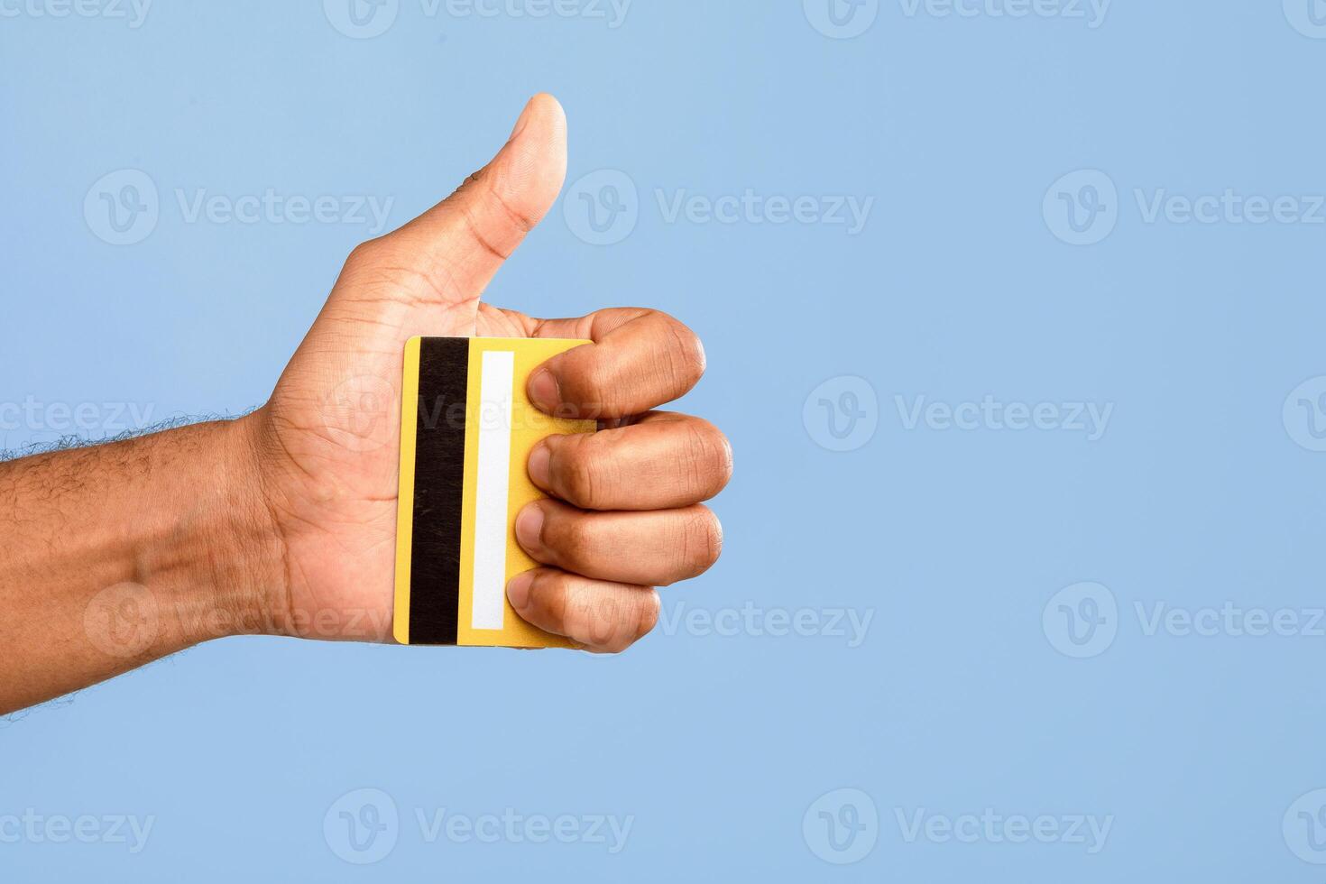 An African American man smiles and shows a thumbs up gesture while holding a yellow credit card in his hand. The background is a solid light blue, suggesting a positive and friendly vibe. photo