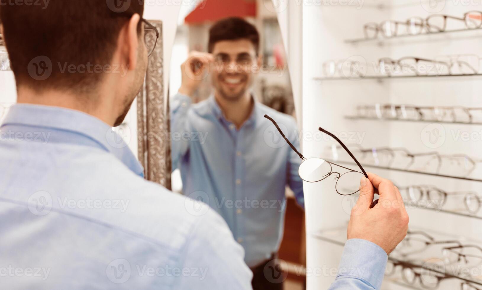 A man stands in an optical store, examining his reflection while trying on a pair of glasses. His smile reflects satisfaction as he considers his new look. photo