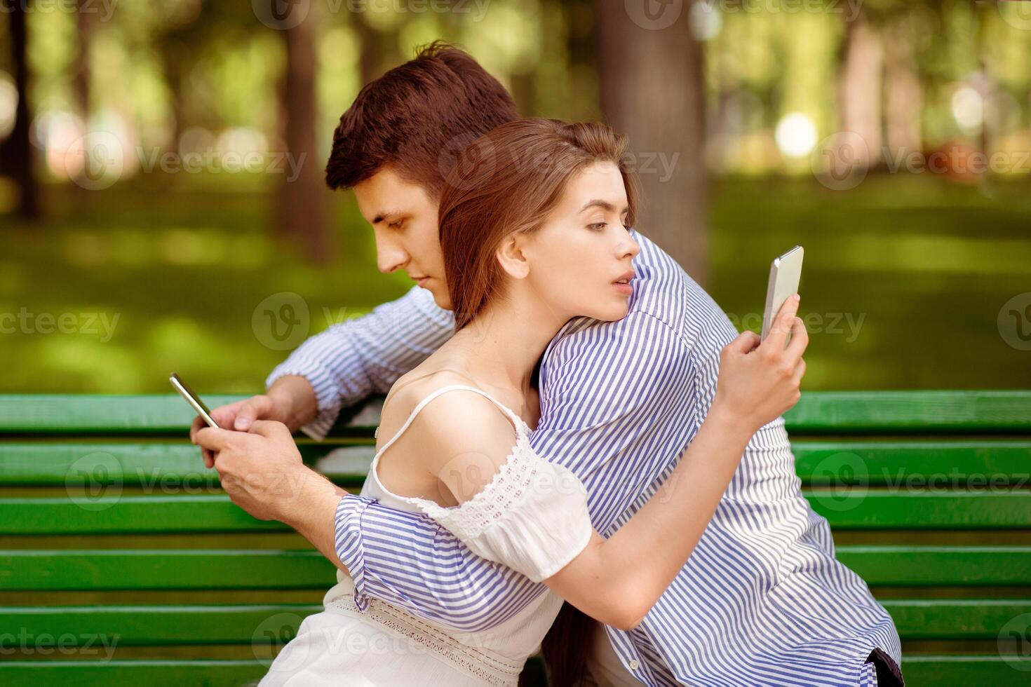 Gadget addicted couple using mobile phones while hugging on bench at park photo
