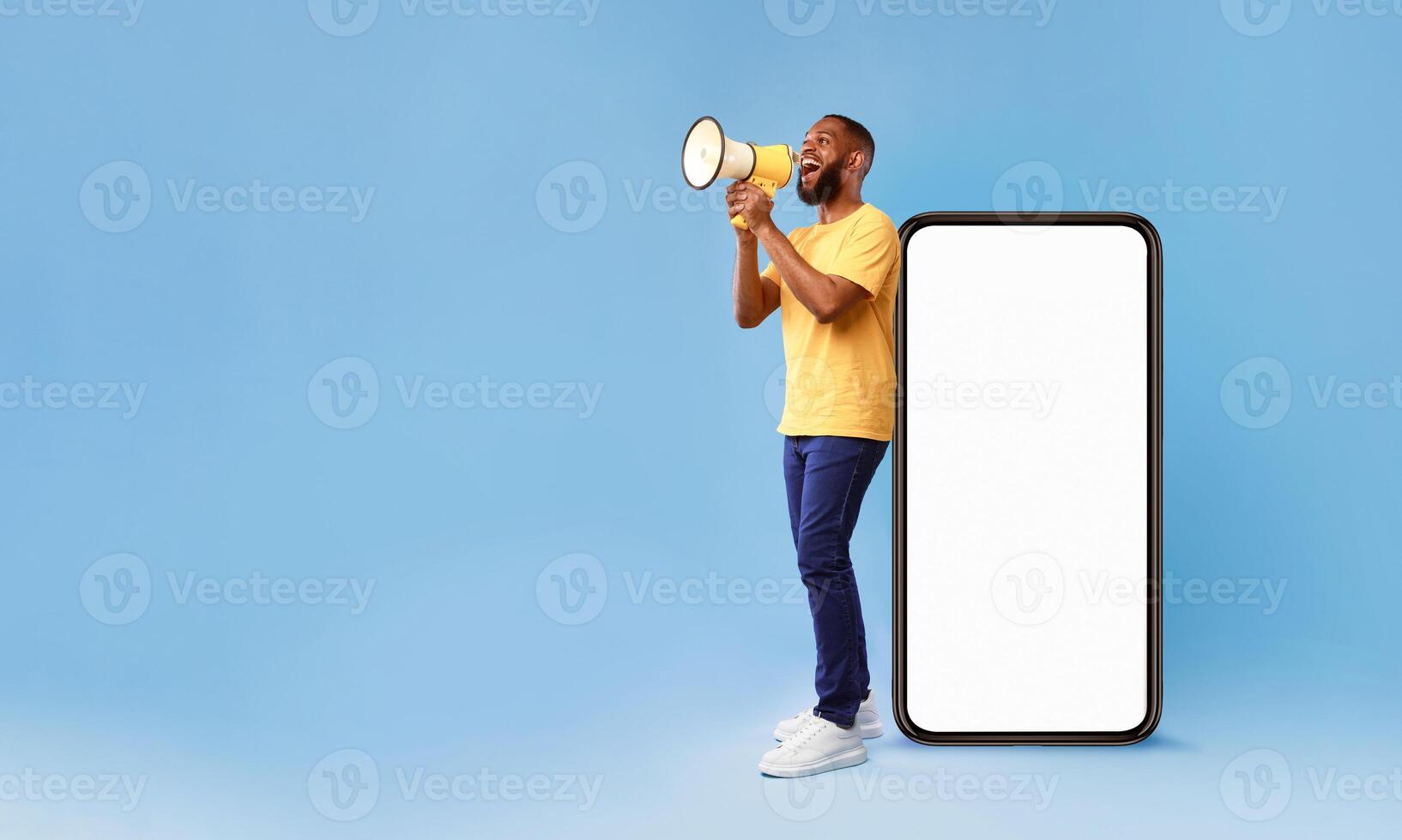 A young African American man enthusiastically holds a megaphone beside a large blank smartphone on a vibrant blue background. His casual outfit and joyful expression convey a lively lifestyle. photo