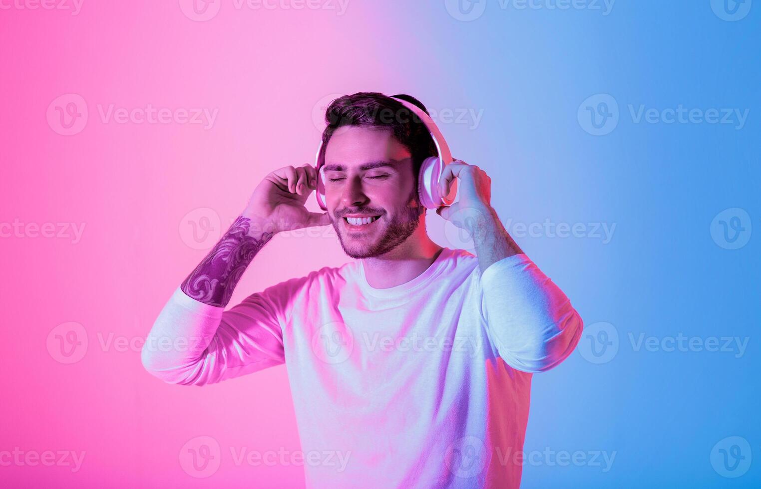 A young man is immersed in music while wearing headphones. He smiles contently with colorful pink and blue lighting creating an energetic atmosphere around him. photo