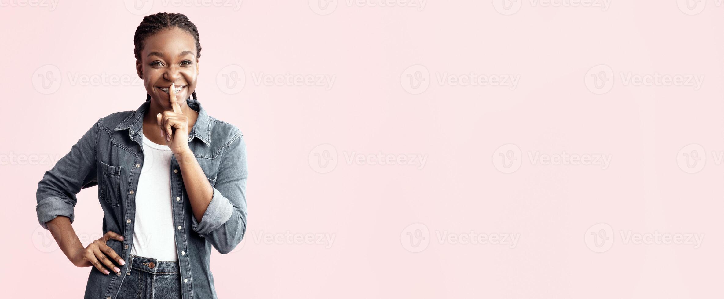 A young woman stands confidently with a smile, playfully placing her finger on her lips. She is dressed casually in a denim jacket and jeans, set against a soft pink backdrop. photo