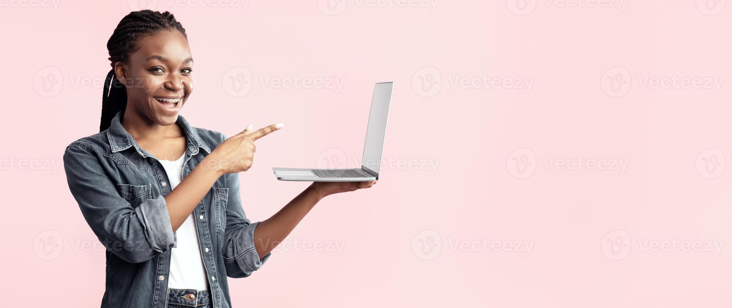 A young woman with braided hair smiles while pointing at her open laptop. She stands against a soft pastel background, appearing enthusiastic about sharing something on the screen. photo