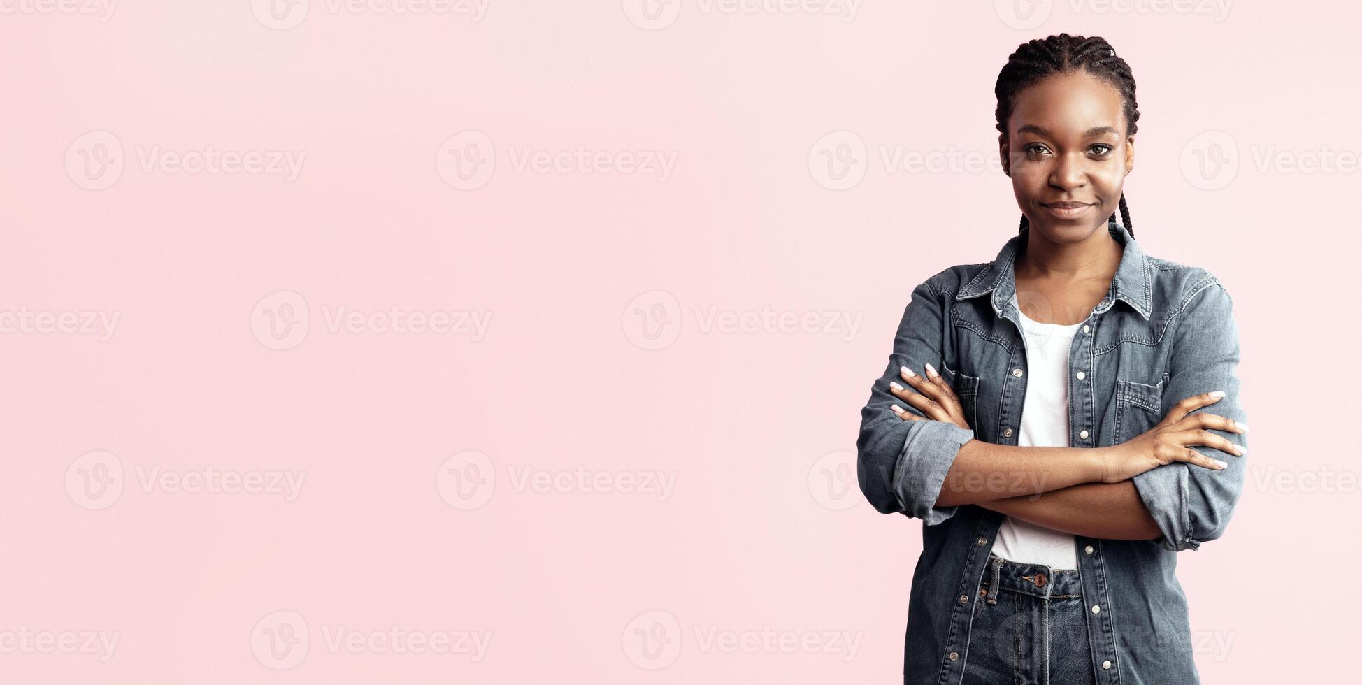 A young woman stands confidently with arms crossed, wearing a denim jacket and white shirt. She has a warm smile and poses against a soft pink backdrop, exuding self-assurance. photo