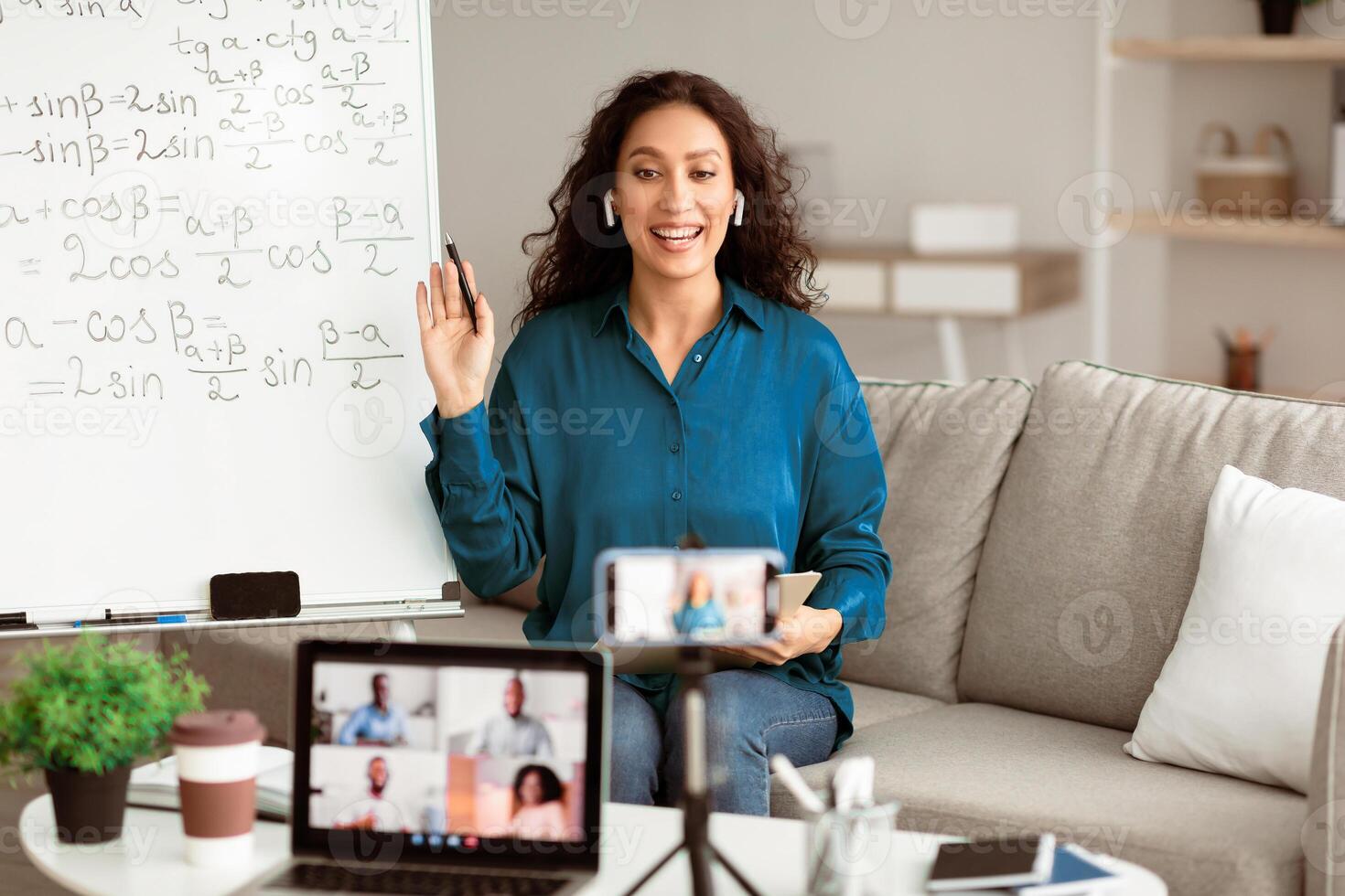 A young Arab woman with brunette hair smiles and gestures while teaching an online class. She is in a cozy living space, with a whiteboard full of equations behind her, engaging with students. photo