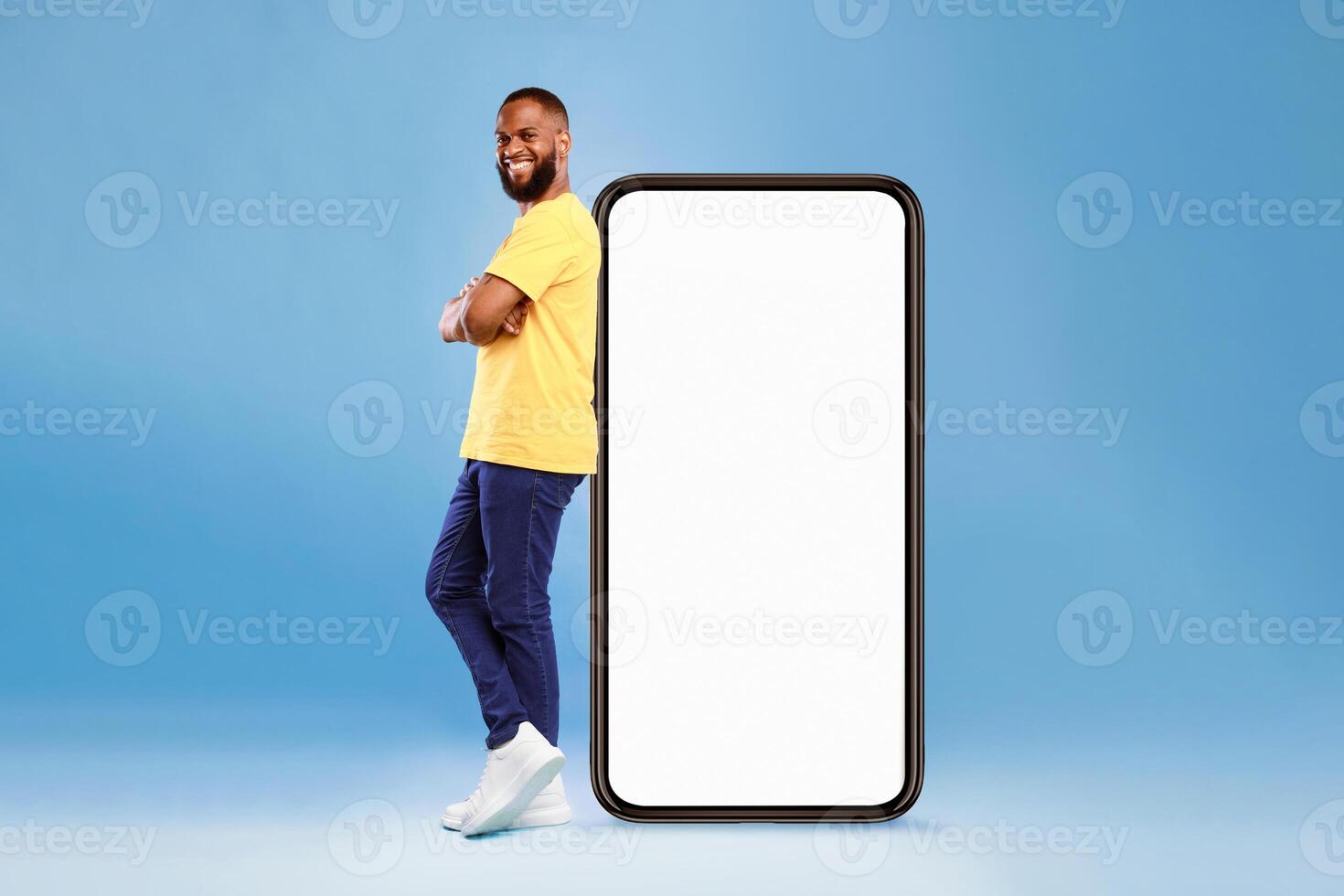 A man with a joyful expression stands confidently next to a large, blank smartphone. He wears a bright yellow shirt and casual pants, showcasing a modern lifestyle in a vibrant studio setting. photo