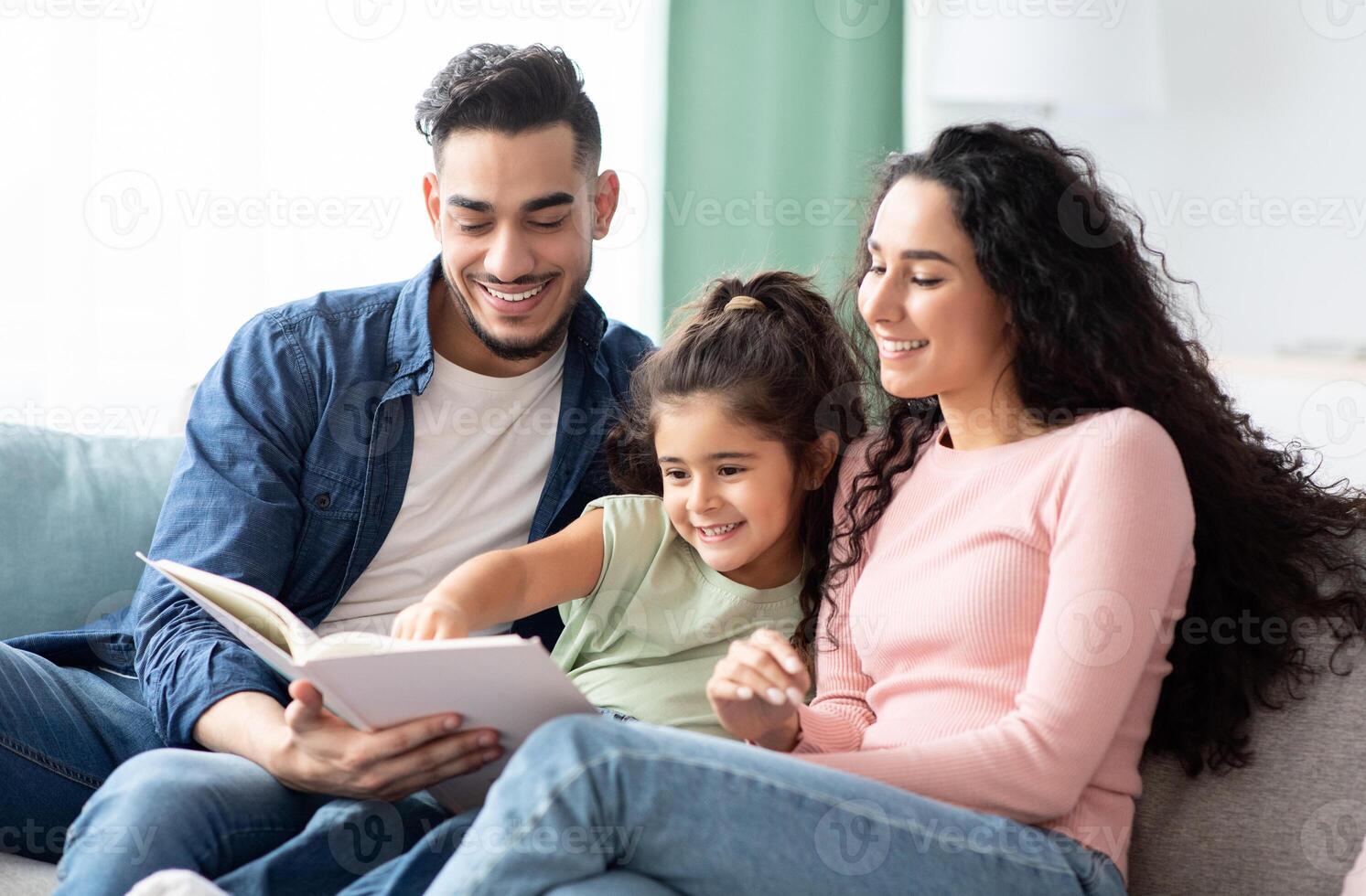 Family Time. Happy Arab Mom, Dad And Little Daughter Reading Book Together While Relaxing On Couch In Living Room, Happy Middle Eastern Parents Having Fun With Their Child At Home, Closeup Shot photo