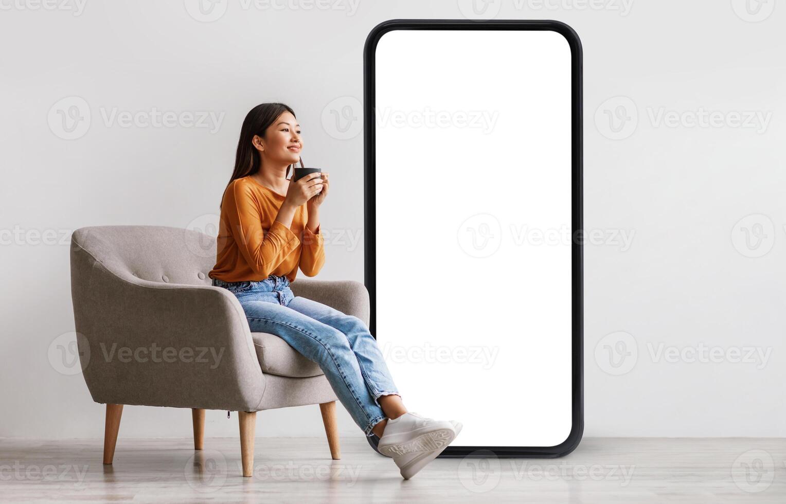 Young Asian woman drinking coffee, having break, relaxing in armchair near giant cellphone with empty screen against white studio wall, mockup for cool new mobile app or online ad design photo