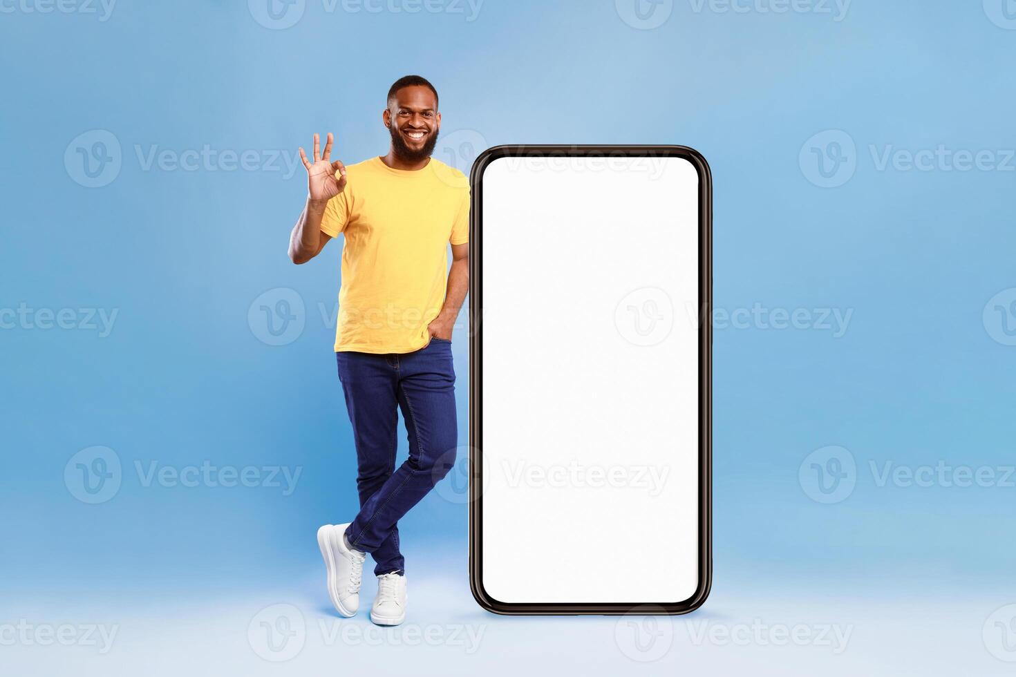 A cheerful African American man, casually dressed in a yellow t-shirt, stands next to a large blank smartphone. He shows a hand sign, exuding positivity against a vibrant blue background. photo