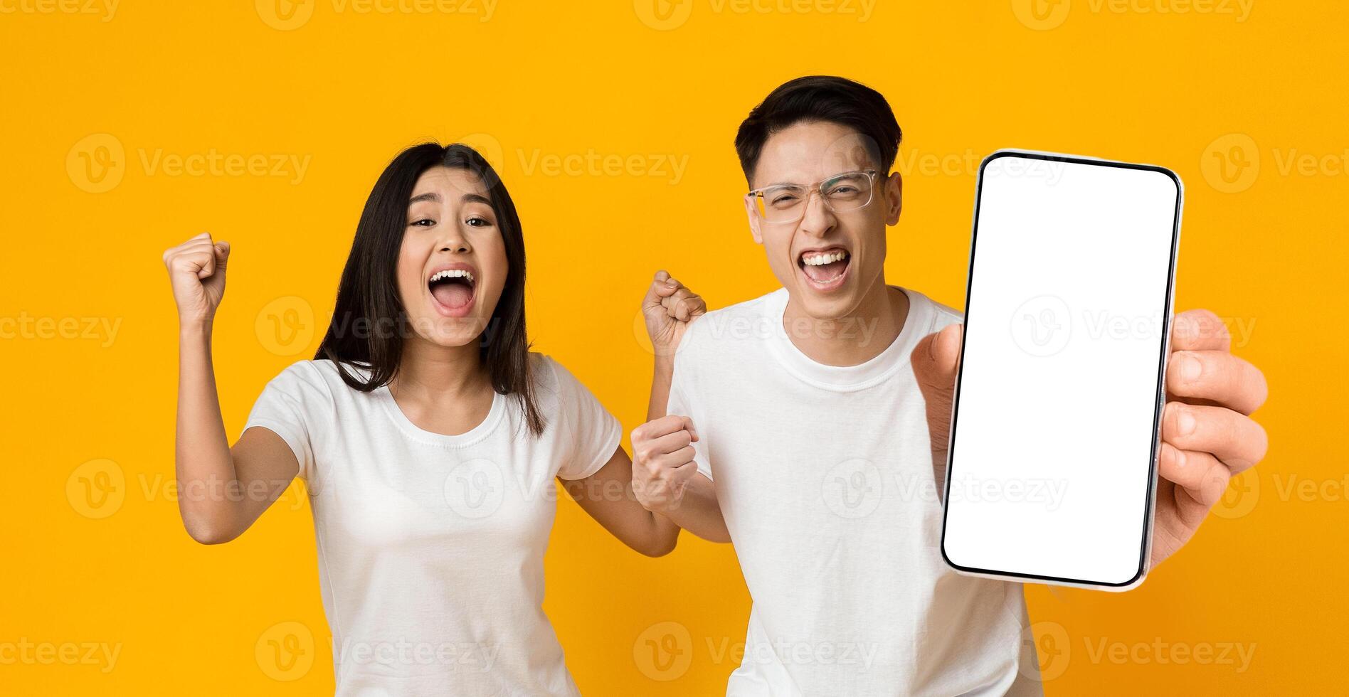 Yes. Excited Asian Couple Holding Big Smartphone With Empty White Cell Screen In Hand Shaking Clenched Fists, Cheerful Guy And Lady Celebrating Win, Yellow Orange Studio Background, Mock Up Copy Space photo