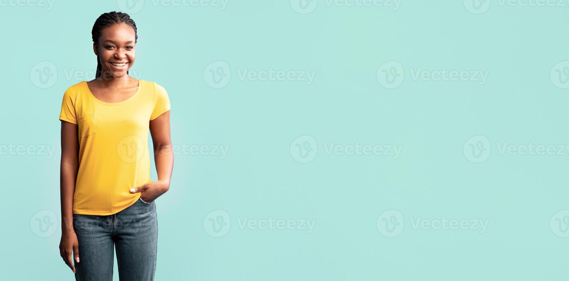 A young woman stands with her hand in her pocket, wearing a yellow shirt and jeans. She smiles warmly, exuding confidence and positivity in front of a soft blue background. photo