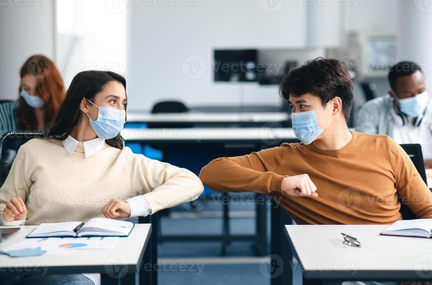 Stop Spreading Virus Concept. Smiling diverse students wearing protective face masks greeting each other and bumping elbows at classroom. Man and woman sitting at desks, studying in high school photo