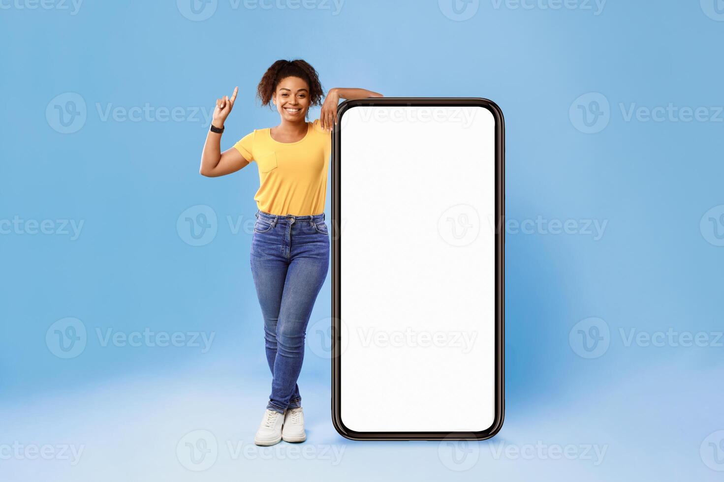 A young African American woman stands with a cheerful expression beside a large blank smartphone, showcasing her style in an isolated setting. This captures a modern lifestyle aesthetic. photo