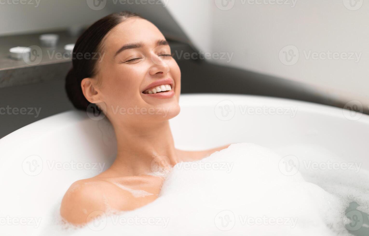 Woman Closing Eyes Taking Bath With Foam Lying In Water And Relaxing In Modern Bathroom Indoors. Gorgeous Young Female Bathing Enjoying Spa Day. Domestic Relaxation, Beauty And Wellness photo