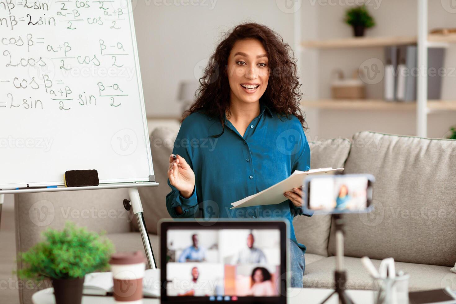 A brunette woman teaches an online class in a well-lit living room. She explains concepts from a whiteboard while engaging with students on her laptop, showcasing an interactive learning environment. photo