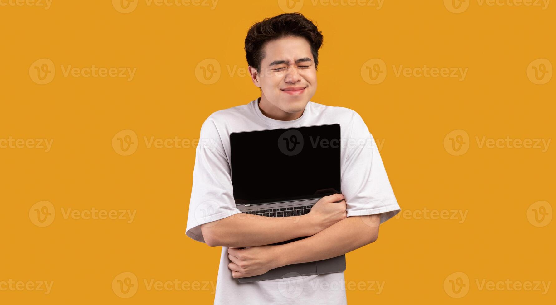 Technology Lover Concept. Happy excited asian man hugging laptop with black blank empty screen, holding it tight near chest. Geek posing isolated over orange studio background wall, copy space photo