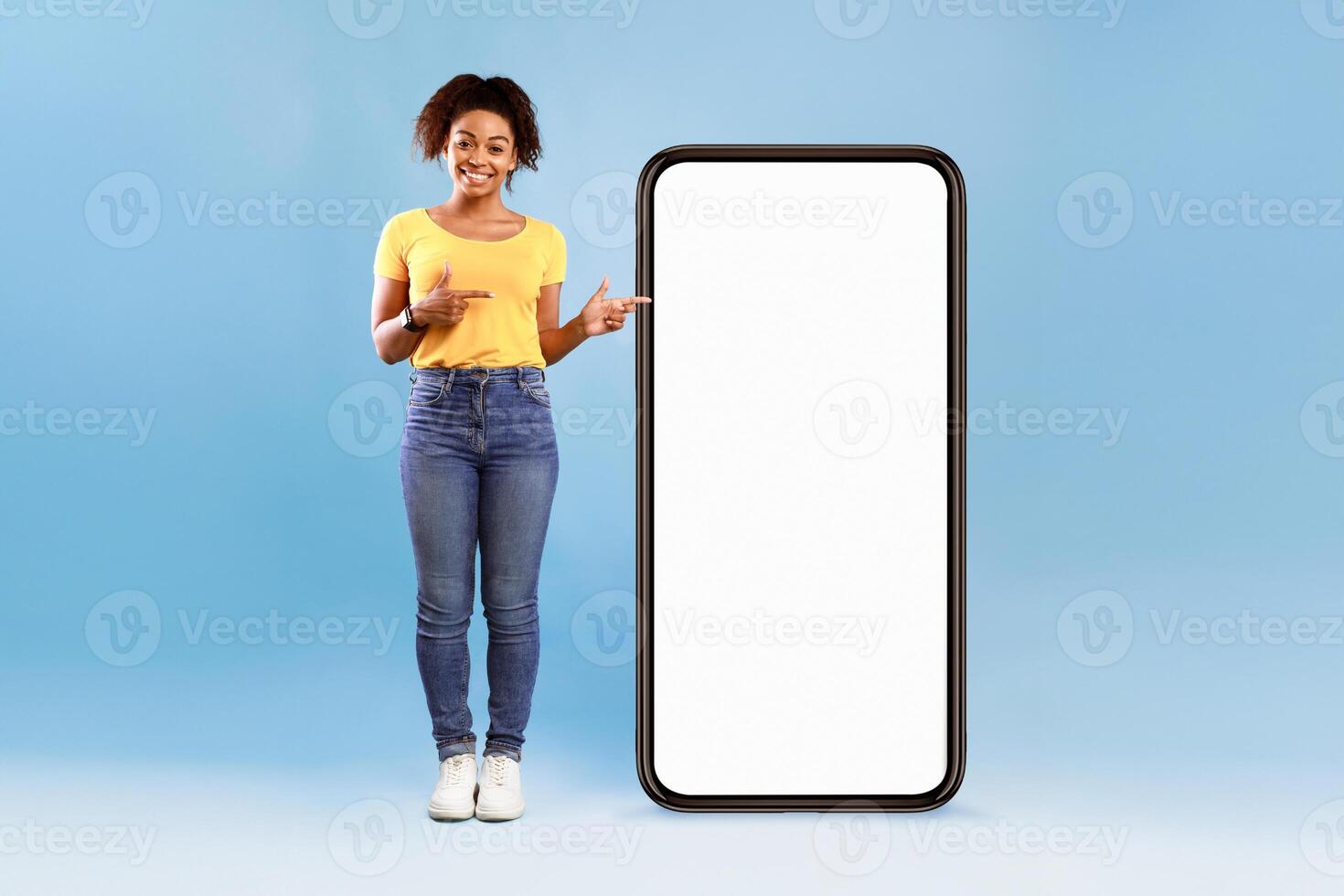 A young African American woman stands against a solid blue background, smiling confidently. She wears a casual yellow shirt and jeans while pointing at a large blank smartphone, inviting attention. photo