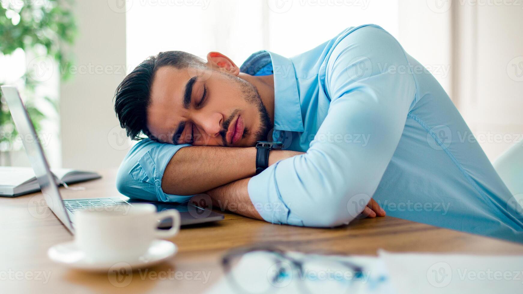 A young Arab man is resting with his head on his arms at a desk in a modern office. The atmosphere indicates a long working day, with a laptop and paperwork nearby. photo
