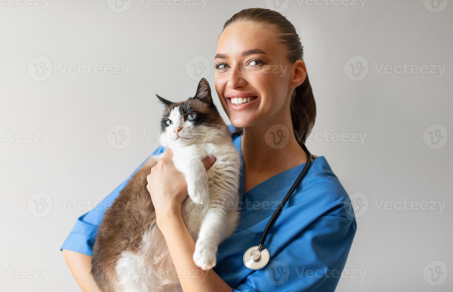 Portrait Of Happy Veterinarian Doctor Woman Holding A Cat, Posing With Healthy Feline Patient Over Gray Studio Background, Smiling At Camera. Pet Health Care And Routine Check Up Concept photo
