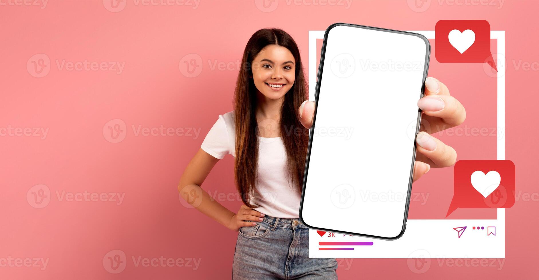 A woman stands against a pink background, smiling while holding up a smartphone. The device displays a blank screen, ideal for showcasing social media applications and engagement. photo