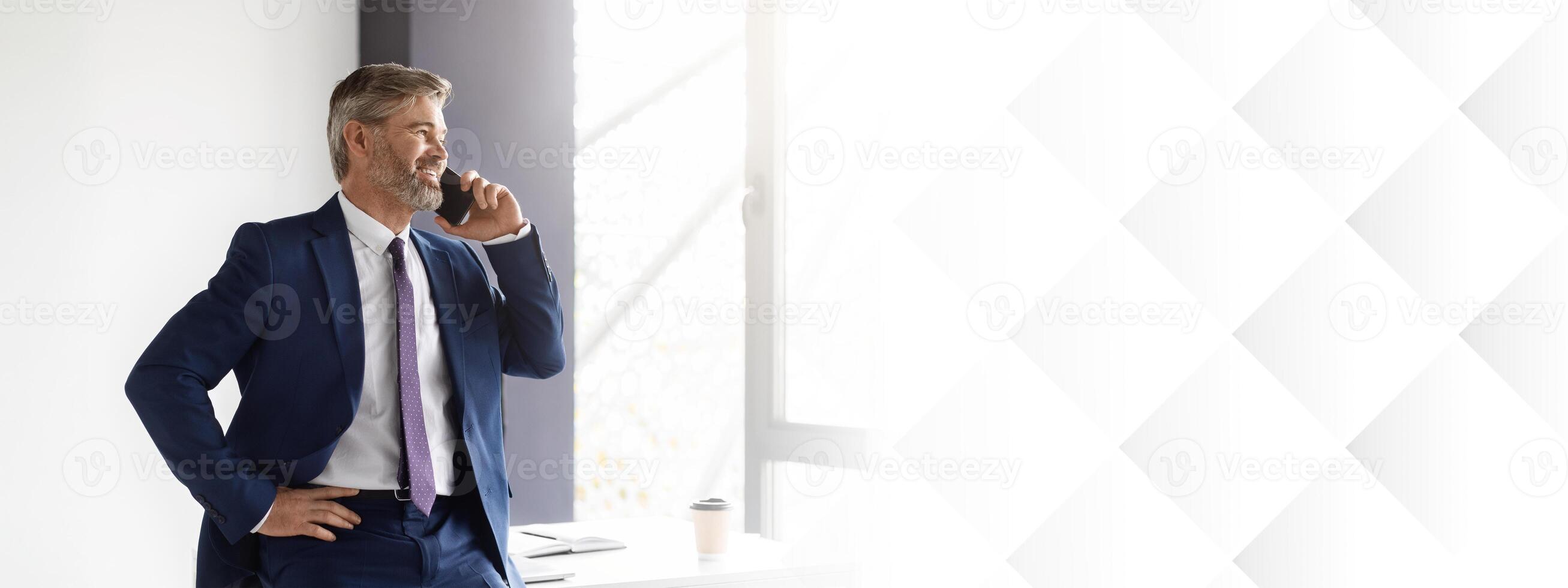 Business Call. Confident Middle Aged Businessman Talking On Cellphone At Office, Handsome Mature Man Entrepreneur Wearing Suit Standing Near Desk And Enjoying Mobile Communication, Copy Space photo