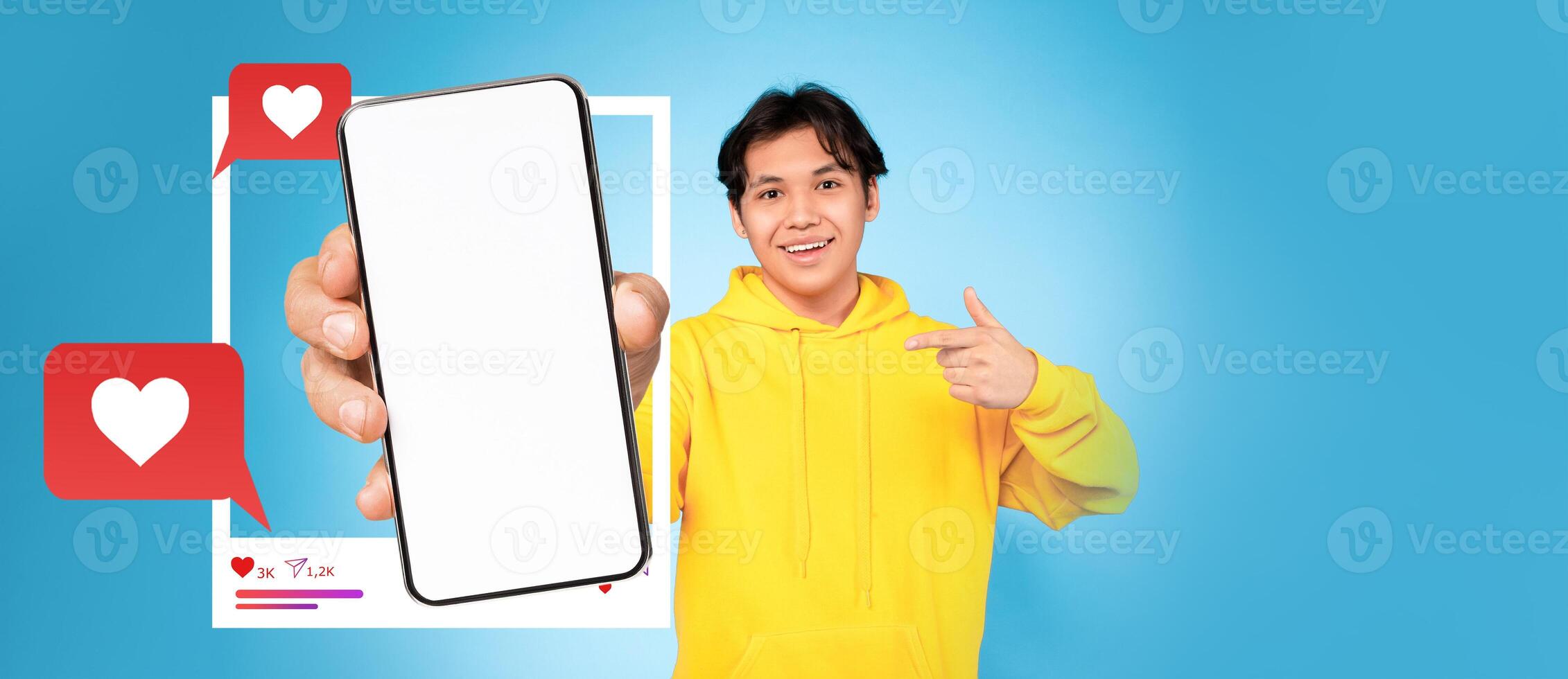 A young influencer in a bright yellow hoodie holds up a smartphone, pointing at the blank screen. The background is a vibrant blue, highlighting social media elements like hearts and chat bubbles. photo