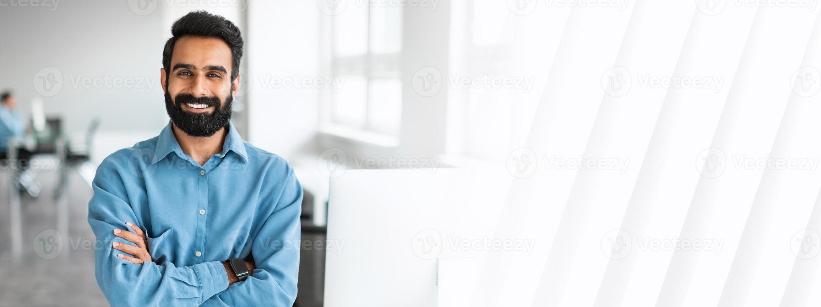 Successful young indian businessman in shirt standing with folded arms near desk, looking at camera and smiling, working in office interior, copy space photo