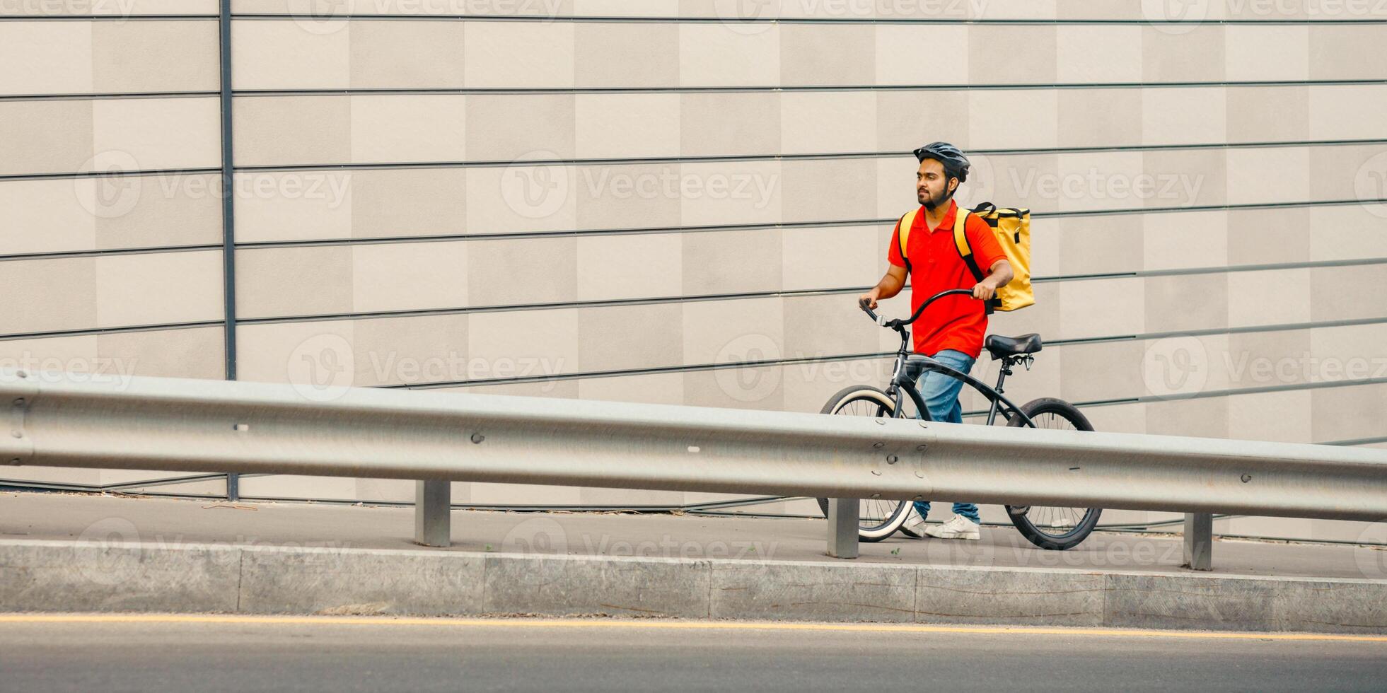 Work deliveryman in city. Courier in safety helmet with yellow backpack and bicycle is tired and walking up street on gray wall background, free space photo