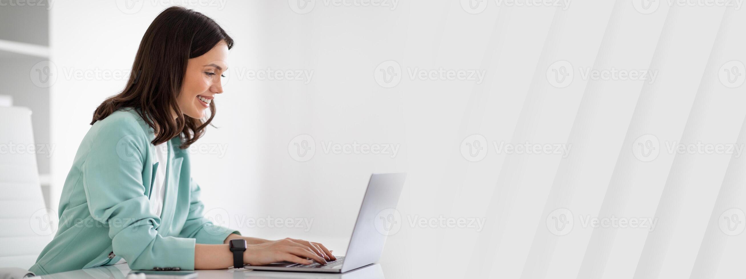Smiling young european lady in suit at table typing on laptop, chatting with client in modern office interior, profile, ad, offer. Businesswoman and business, occupation, work, lifestyle with device photo