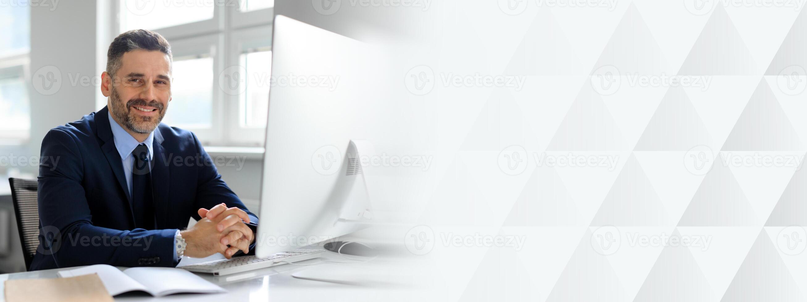 A mature Caucasian businessman is seated at his office desk, exuding confidence and professionalism. He smiles as he engages with his work, surrounded by a bright and organized workspace, copy space photo