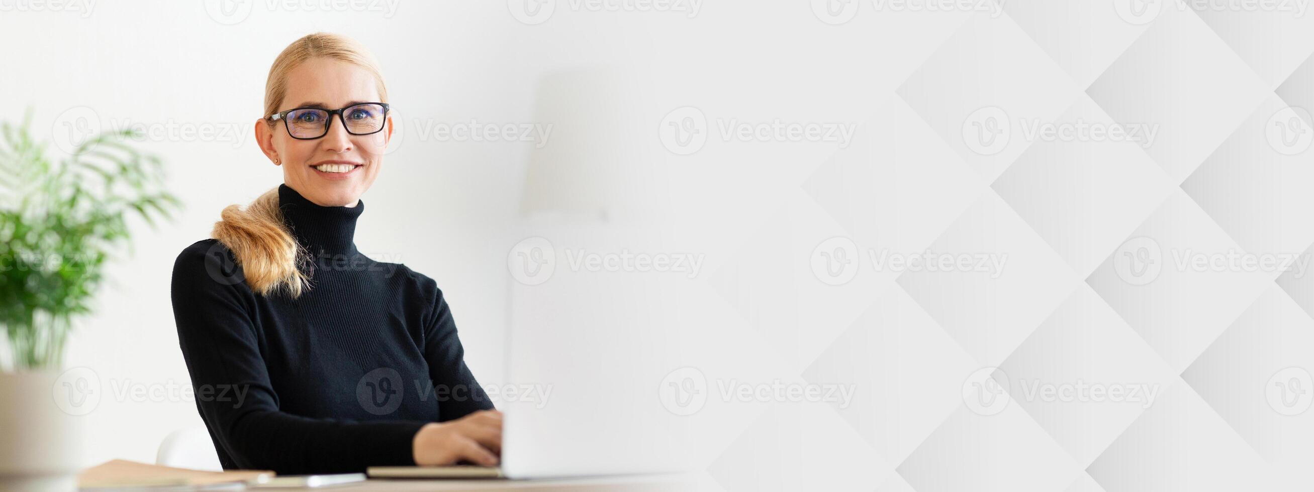 A woman with glasses smiles while sitting at her desk, engaging with a laptop. The modern office features greenery and a neutral color palette, creating a bright and inviting atmosphere, copy space photo