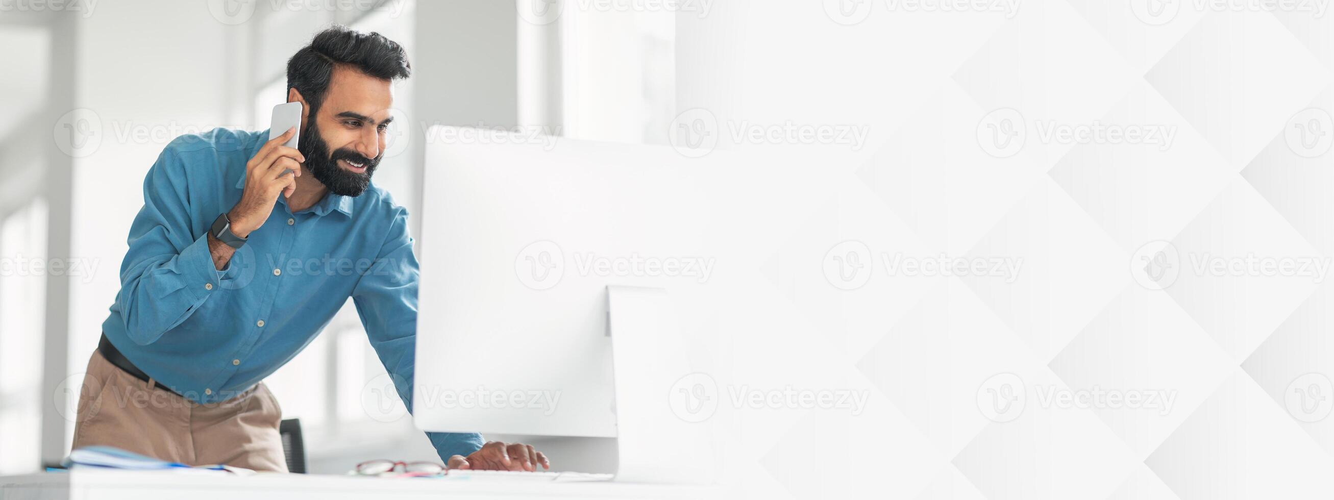 Cheerful bearded indian man in blue shirt multitasking, talking on phone and looking at computer monitor, working in light-filled contemporary office interior photo