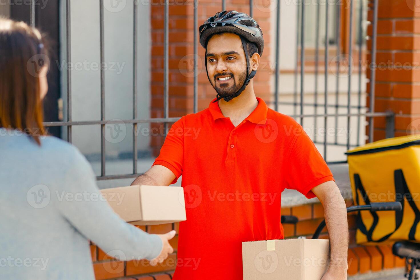 Fast eco service and delivery of parcels to home. Indian smiling millennial man in orange t-shirt and safety helmet gives to lady client a box with parcel, next to bicycle with yellow thermo bag photo