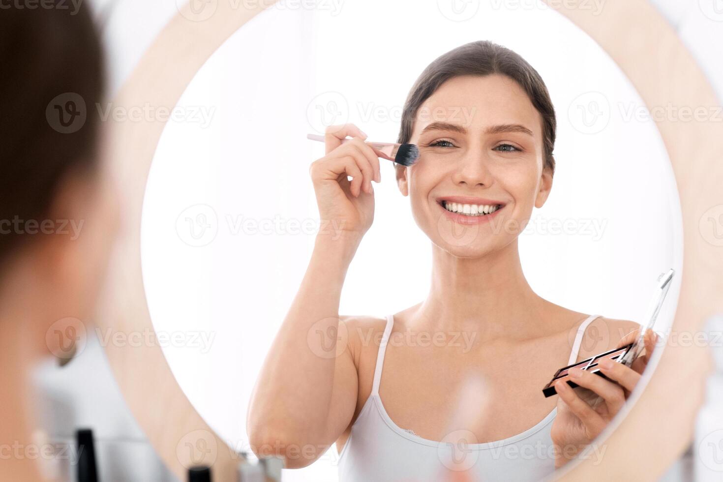 Smiling attractive young lady doing makeup before going out, applying blush on her cheeks. Beautiful cheerful woman using beauty products at home, putting highlighter with brush on her face photo