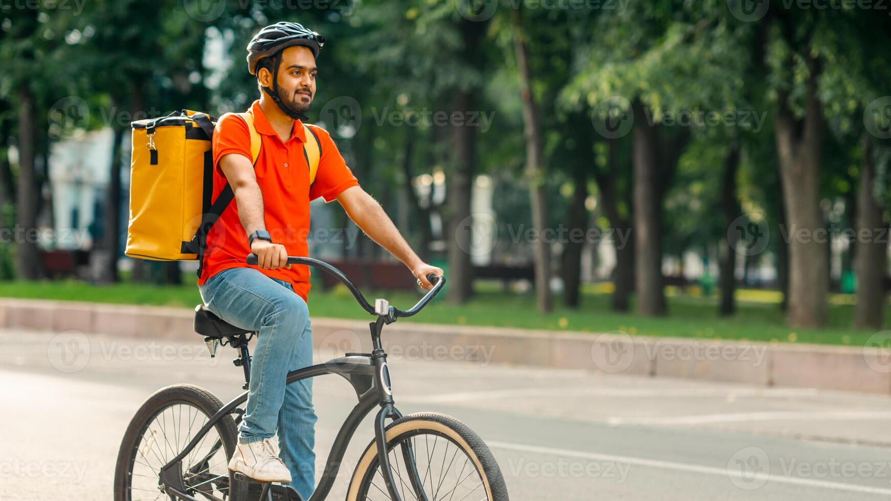 Modern work and delivery on eco transport. Young friendly indian man in uniform, protective gear with backpack riding on bicycle on sidewalk down the street, side view, panorama, empty space, outdoor photo