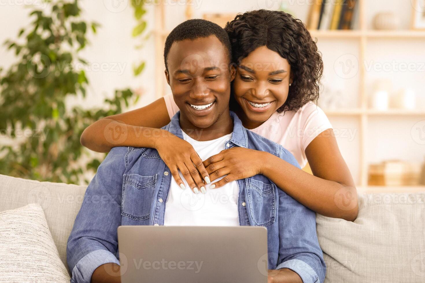 Loving african american couple using laptop while resting at home, watching photos together, copy space. Happy black family husband and wife spending time together at living room, woman hugging man