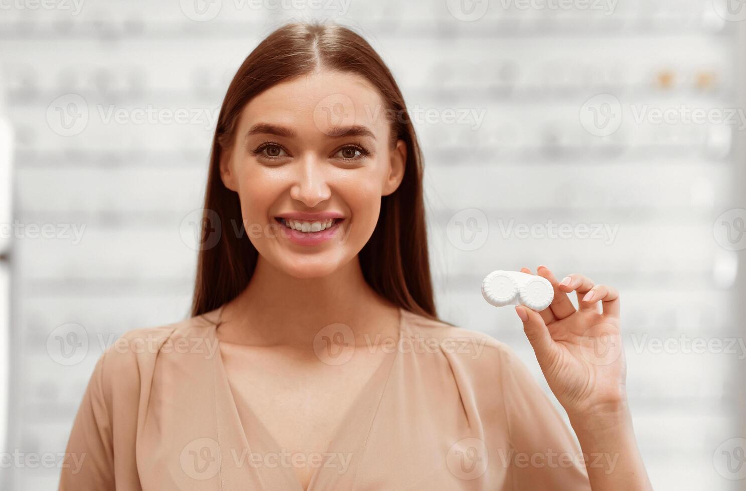 A young woman smiles while holding a contact lens case in an eyewear store. The store has various eyeglasses displayed in the background, indicating a vibrant shopping atmosphere. photo