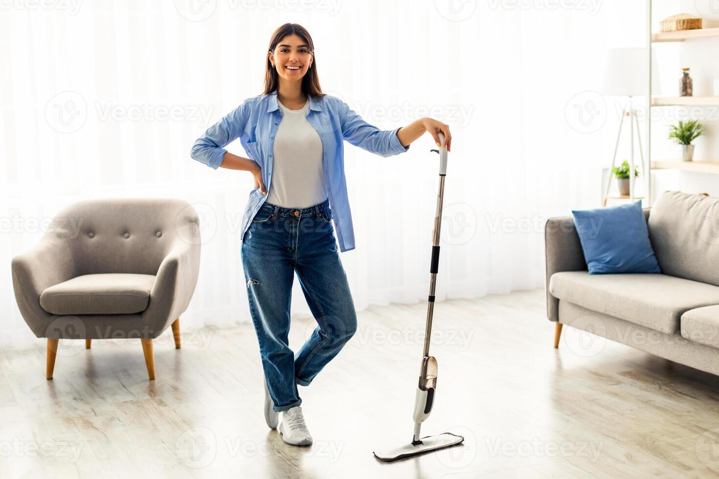 A young woman is happily engaging in house cleaning. Dressed casually, she stands with a cleaning tool in her modern living room, creating a fresh atmosphere in her space. photo