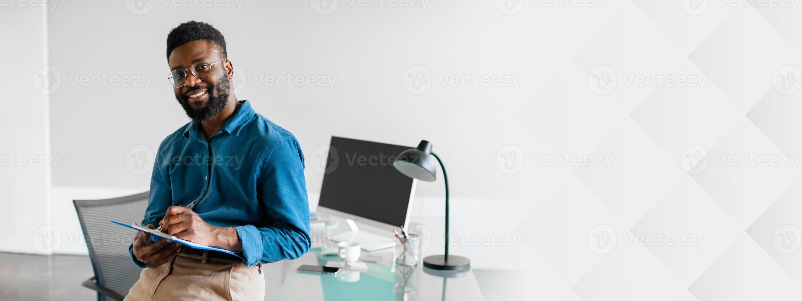 In a contemporary office space, a young African American businessman sits confidently with a clipboard. He smiles while showcasing a professional and focused approach to his work, copy space photo