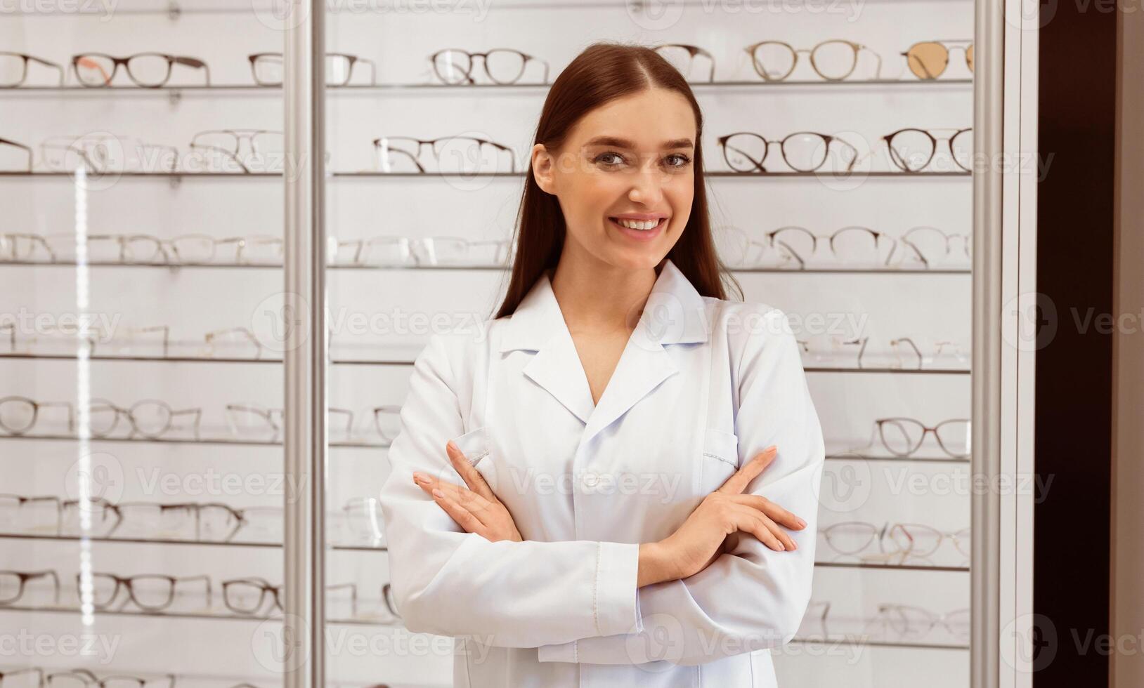 A smiling professional stands confidently with arms crossed in front of a display of various eyeglasses. The store's organized layout highlights the diversity of available frames. photo