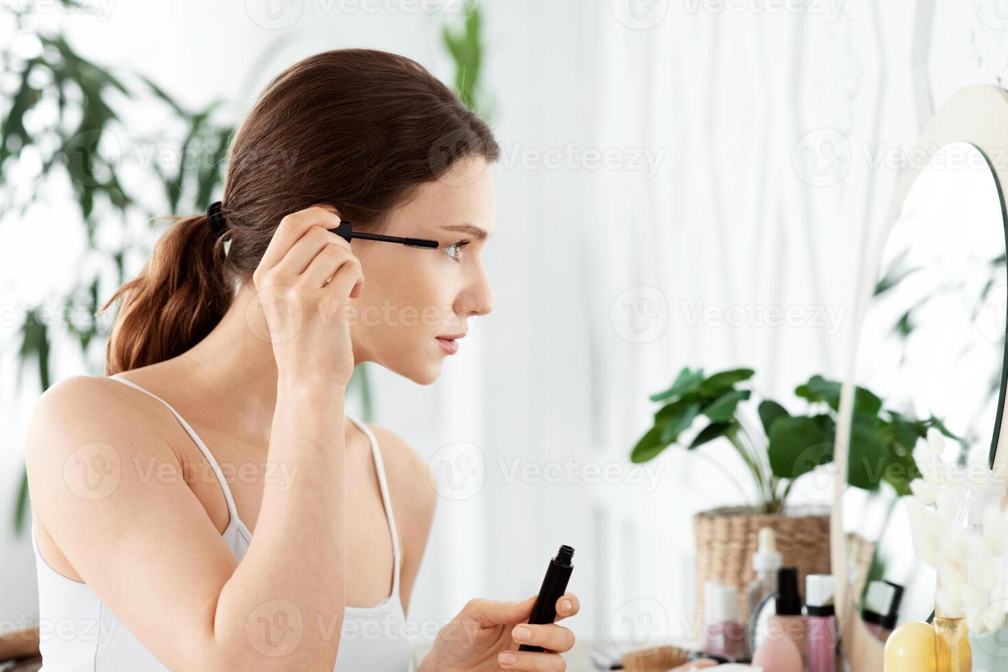 Side view of young woman looking at mirror, applying mascara, home interior, copy space. Cheerful attractive lady in homewear sitting in bedroom, using mascara, getting ready before going out photo