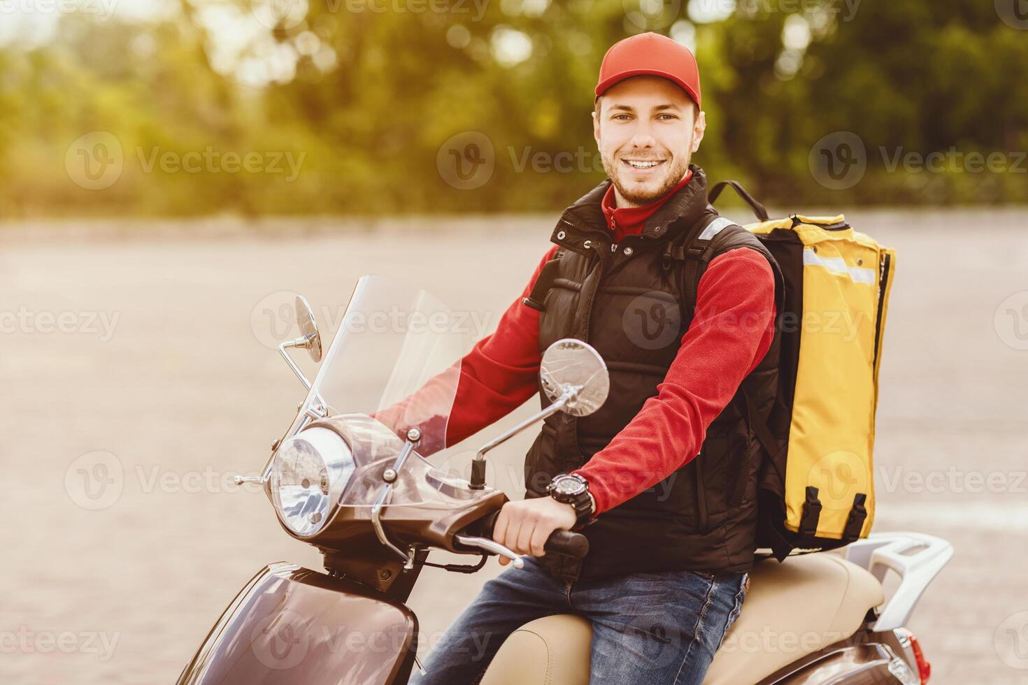 Cheerful Courier Sitting On Scooter Delivering Food In Yellow Backpack Bag, Posing Smiling To Camera Outdoors. Empty Space photo