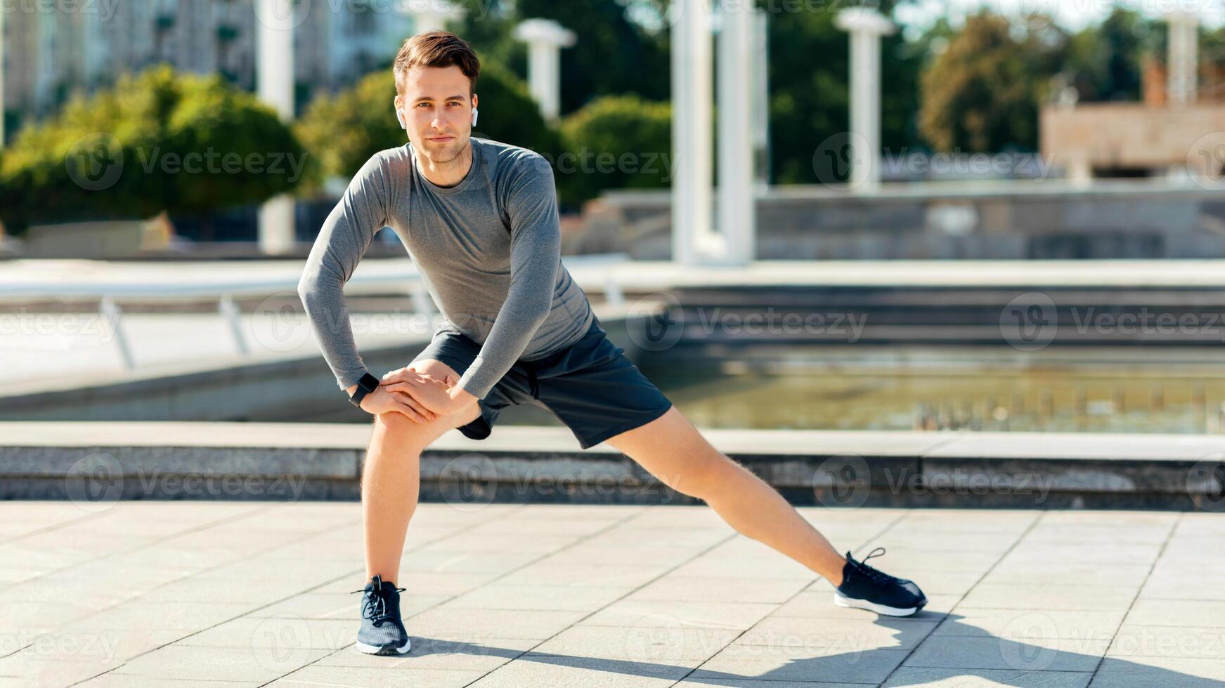 Training before marathon. Young man with wireless headphones in sport uniform doing exercises for legs, free space photo