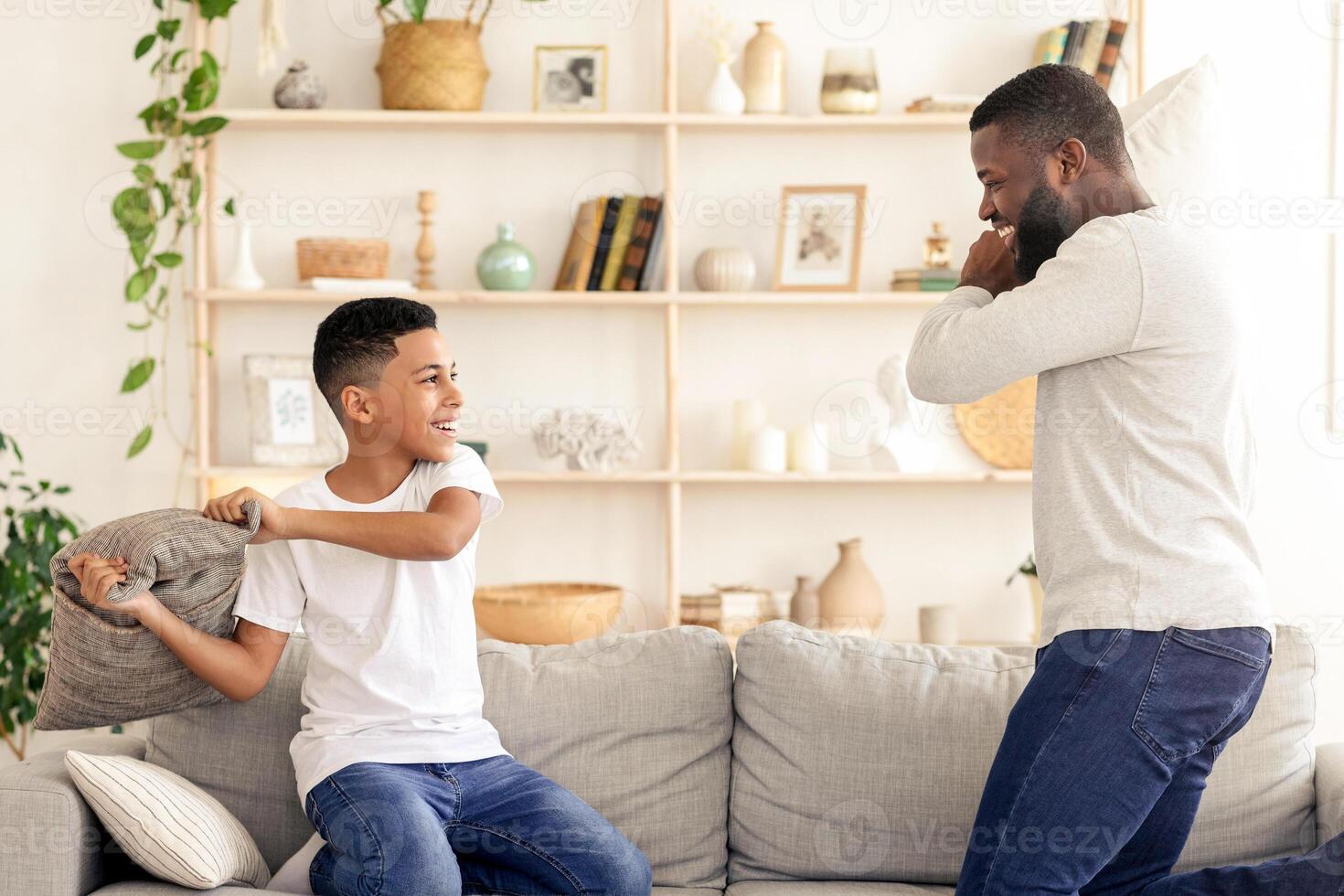 A young boy smiles as he prepares to hit his father with a pillow during their playful tussle on the couch. The cozy living room is adorned with plants and decorative items. photo