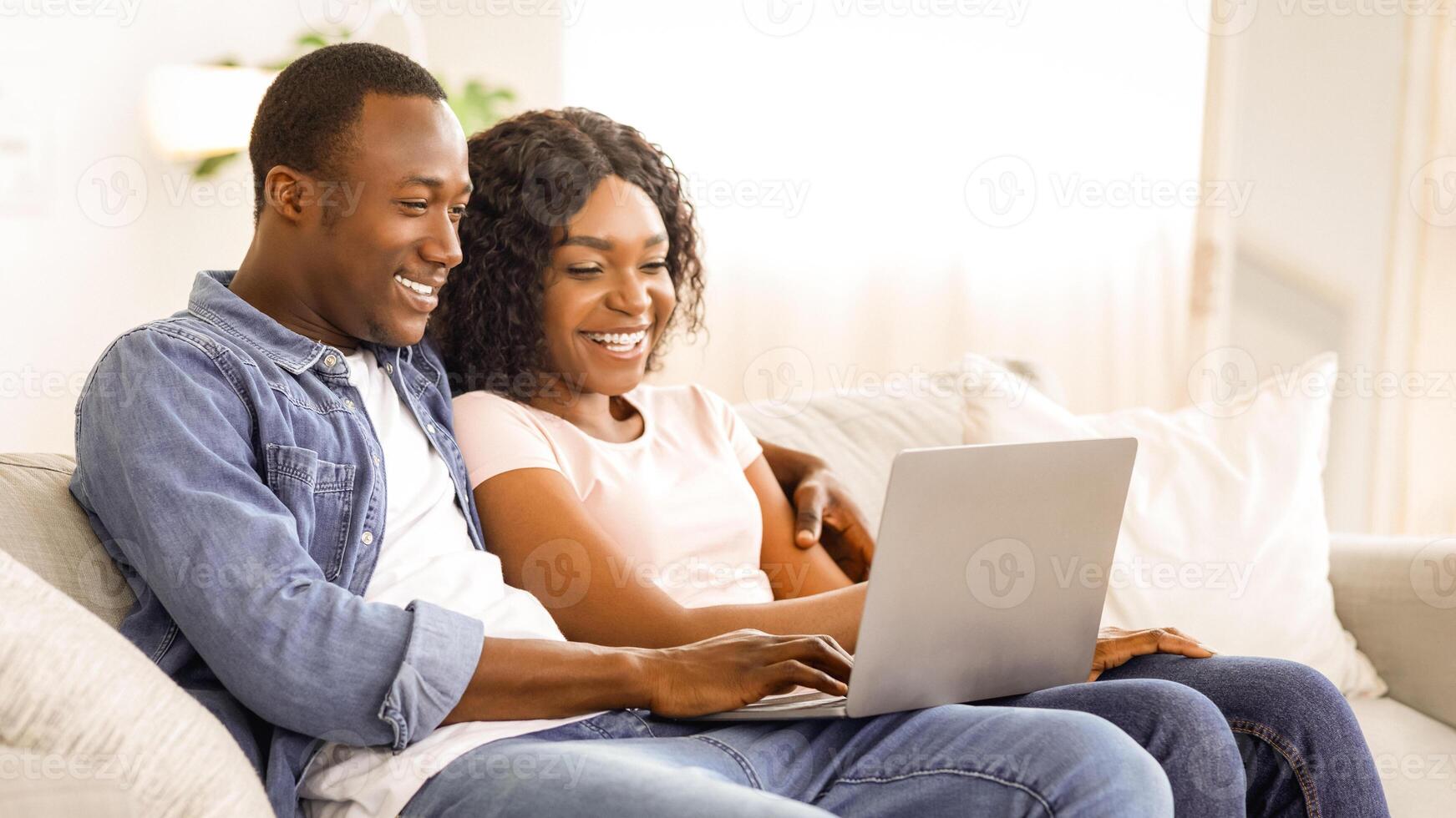 Cheerful black couple using laptop while resting on couch at living room at home, watching funny content together, copy space photo