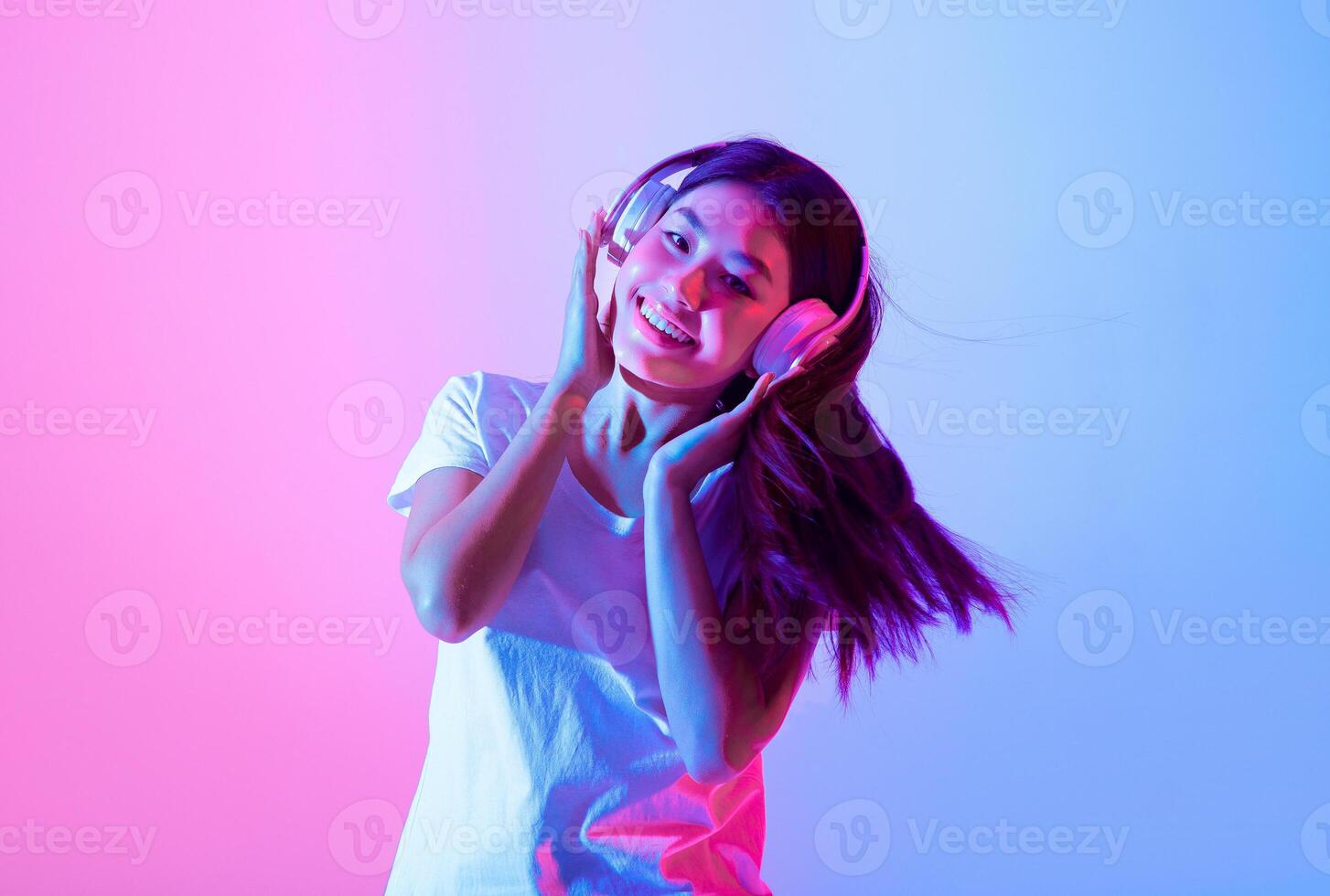 A young Asian woman with long hair smiles while wearing headphones. Colorful pink and blue lighting creates a vibrant atmosphere as she enjoys her music, embodying joy and relaxation. photo
